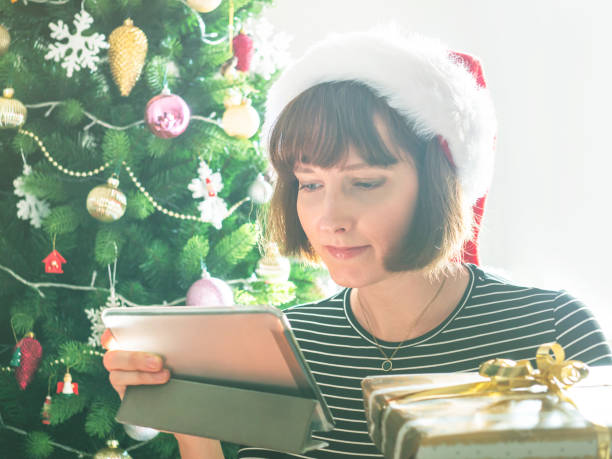 istock Smiling woman holding wrapped gifts using tablet near christmas tree 1420855880