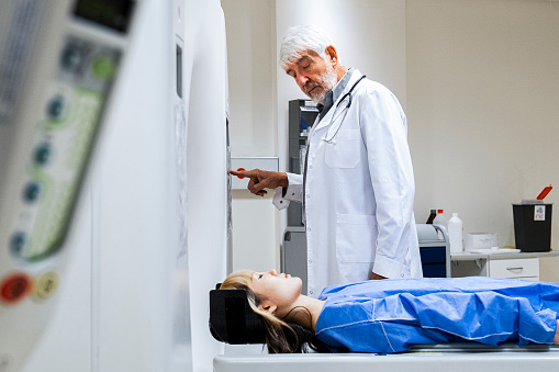 Mid-shot side view of senior doctor explaining MRI scan procedure to female patient laying on scanner bed.