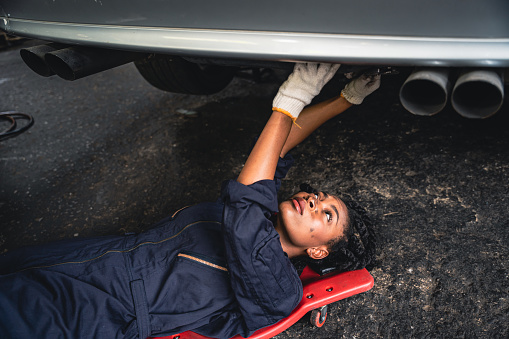 Professional female mechanic lying on slide board and checking the car suspension in the garage.
