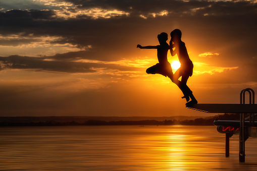 silhouettes of two unrecognizable boys - jumping into the water at sunset - part of a series