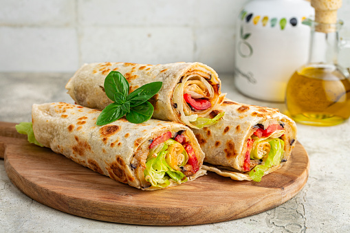 Wraps with a soft flatbread rolled around a filling with vegetables and cheese, tomato, fried eggplant, lettuce. Vegetarian food concept. Light background.