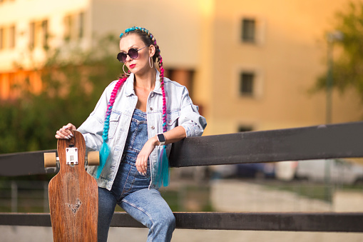 Attractive Caucasian Female Dressed in Trendy Stylish Streetwear With Colorful Hairtails And Glasses Resting With Longboard in City As Recreation Chill At Sunset.Horizontal Image