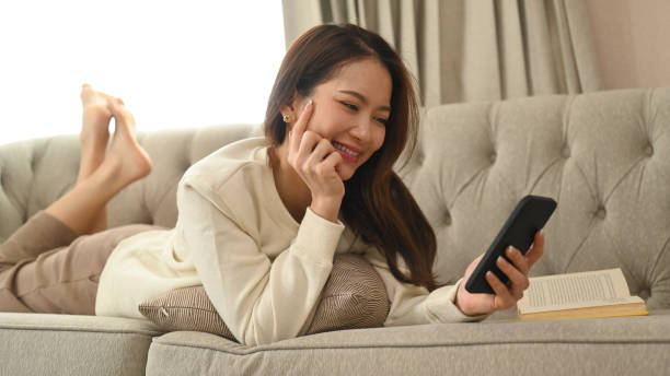 istock Cheerful young woman in warm sweater lying on couch and using mobile phone, spending time in social media internet. 1420927010