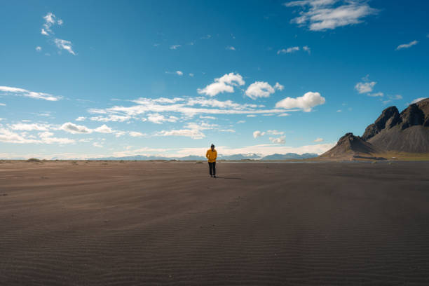 Young asian woman in yellow jacket walking on black sand beach in sunny day at Vestrahorn, Iceland stock photo