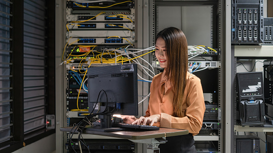 A female programmer is working in a server room. The girl is standing next to the computer racks of the data center. The administrator looks at the monitor with network settings.