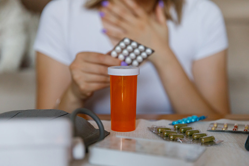Close up shot of an empty pill bottle resting on a table with unrecognizable young woman holding a hand on chest and a medicine packet. Part of a series.