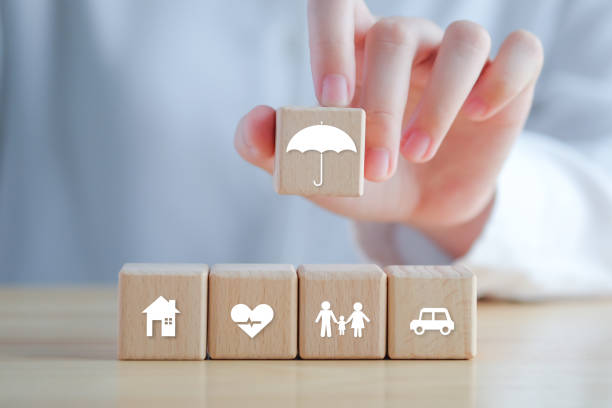 보험 개념. 가능한 만일의 사태에 대한 보호. 보증 생활 개념을위한 나무 블록에 우산 아이콘과 하우스, 자동차, 가족 및 건강 아이콘을 들고 손. - insurance company 뉴스 사진 이미지
