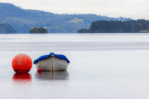 Empty rowing boat floating on lake at Loch Lomond Scotland. Calm serene landscape image of a wooden boat with blue cover moored next to an orange buoy in open water with mountainous background.