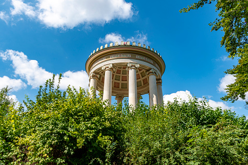 The dome and columns of the Monopteros Temple in the English Garden in Munich from below, with blue skies and light clouds with the foliage of trees in the foreground