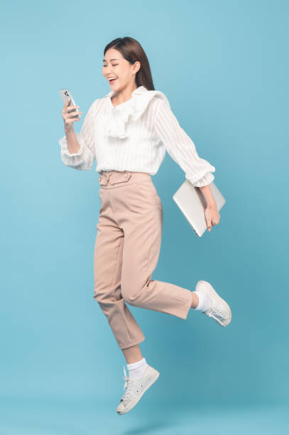 Young beautiful asian woman with smart casual cloth use smartphone and jump isolated on blue background stock photo