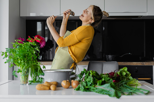 Active 30 years woman housewife holding ladle like microphone singing dancing in modern cozy kitchen at home near vegetables on table. Hobby and karaoke, lifestyle concept. Humor, power in vegetables