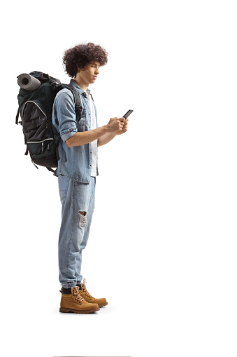 Full length profile of a young male backpacker standing and using a smartphone isolated on white background