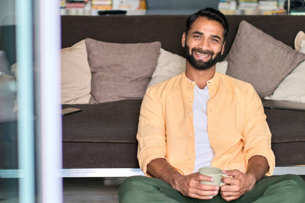 istock Happy indian man looking at camera drinking coffee sitting on sofa at home. 1420975009