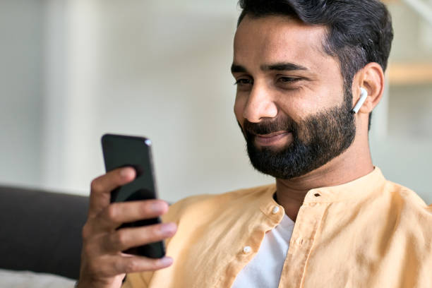 istock Smiling indian man sitting at home wearing earphones using smartphone. 1420975021