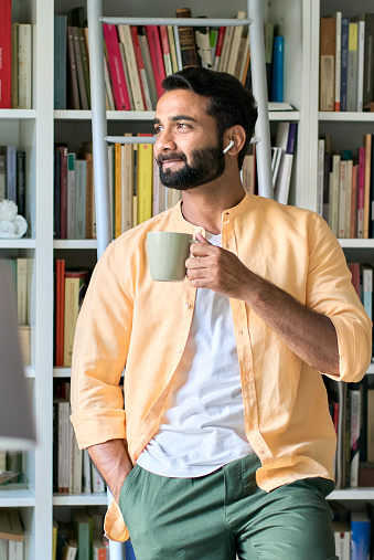 Thoughtful indian eastern ethnic man user standing at home in front of bookcase wearing earbuds drinking coffee  thinking looking through window listening music, audio book or podcast.