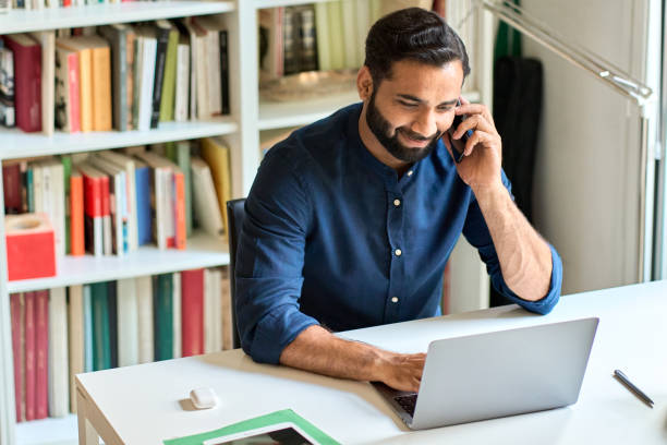istock Smiling indian business man sitting at work desk talking on phone making call. 1420975048