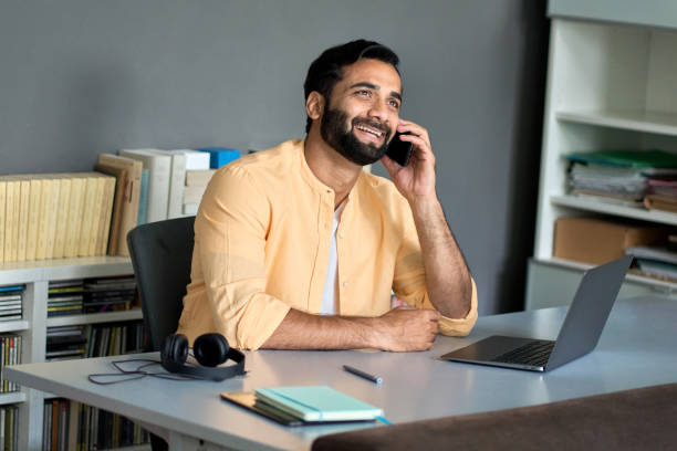 istock Happy indian business man sitting at work desk talking on phone making call. 1420975104