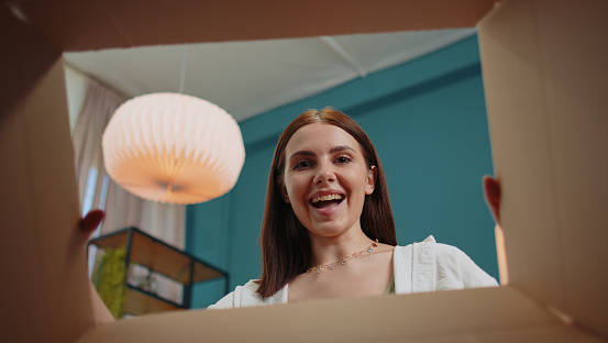 Happy surprised winner girl unpacking delivery parcel sitting at home. Smiling satisfied young woman shopper, online shop customer opening cardboard box receiving purchase gift by fast postal shipping