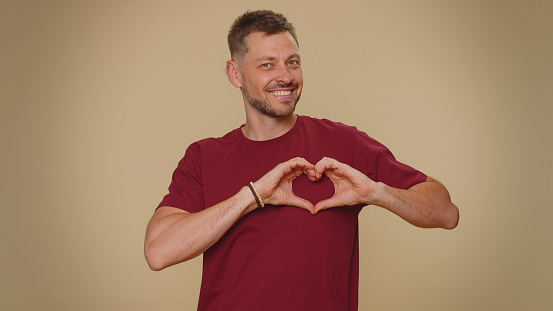 I love you. Smiling bearded handsome young man makes heart gesture demonstrates love sign expresses good feelings and sympathy. Adult stylish male guy isolated on beige studio background indoors