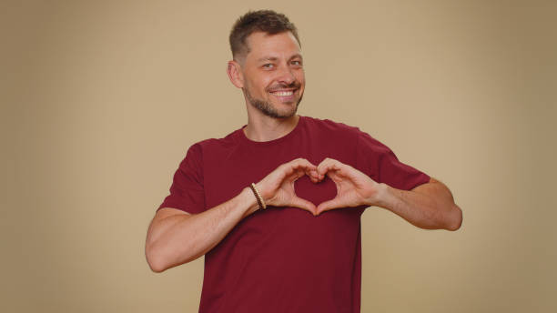 Smiling bearded handsome young man makes heart gesture demonstrate love sign expresses good feelings I love you. Smiling bearded handsome young man makes heart gesture demonstrates love sign expresses good feelings and sympathy. Adult stylish male guy isolated on beige studio background indoors man showing peace sign stock pictures, royalty-free photos & images