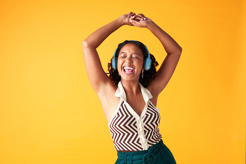 Dance, music and creative woman listening with wireless headphones in photographer studio background. Happiness, freedom and relax female singing while streaming radio or spotify in yellow backdrop
