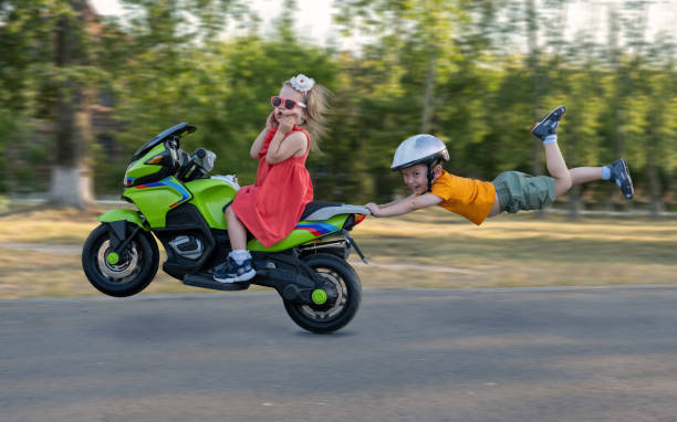 children ride in the park on a children's electric car. a comic plot in which a boy holds onto the seat of a motorcycle and flies in the air surprises the girl. - two wheel imagens e fotografias de stock