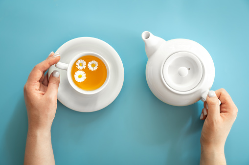 A cup of chamomile tea and a teapot in female hands on a blue background, flat lay, minimalism.