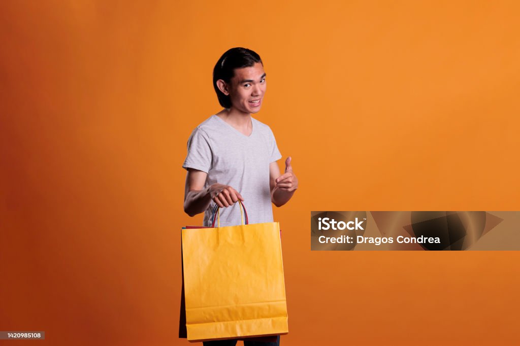 Cheerful smiling man pointing at paper shopping bag Cheerful smiling man pointing at paper shopping bag, store sale advertisement. Happy buyer with purchase, shopaholic buying products, young asian shop customer holding package Adults Only Stock Photo