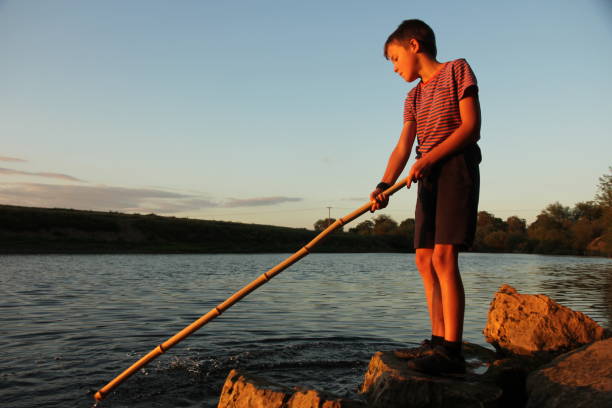 Young boy standing next to the river at sunset playing with a stick and splashing in water