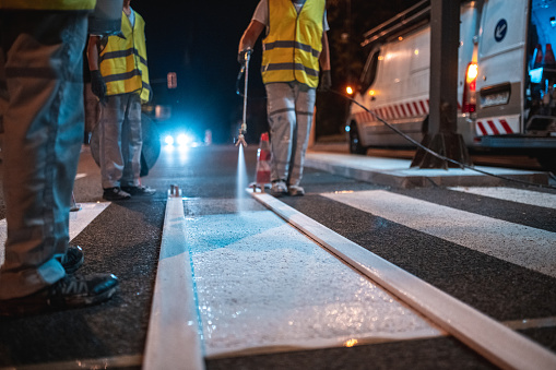 Construction worker wearing a reflective vest spray painting a crosswalk with a new layer of white paint using a spray gun.