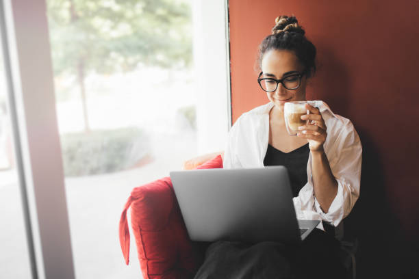 istock Young woman using laptop and drinking coffee sitting near window. 1420989771