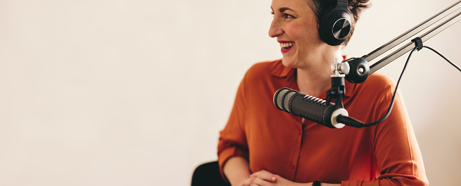 Cheerful woman smiling happily while interviewing a guest on her radio show. Female radio presenter hosting a live audio broadcast in a studio. Woman sitting behind a microphone with a headset.