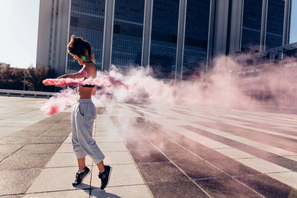 Woman holding colorful smoke grenade outdoors in city. Urban sportswoman with smoke sticks.