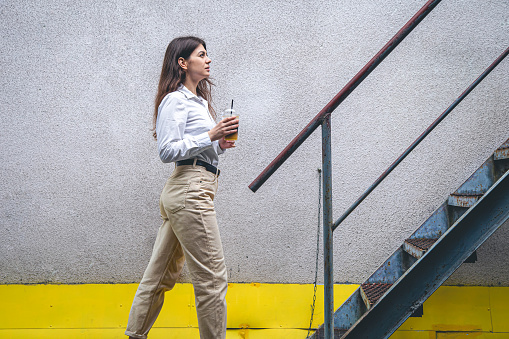 A business young woman in a shirt stands near the stairs with a cup of coffee.