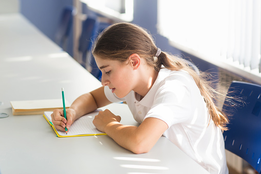 student sits at desk in school classroom and studies. girl writes with pencil in notebook in ruler.