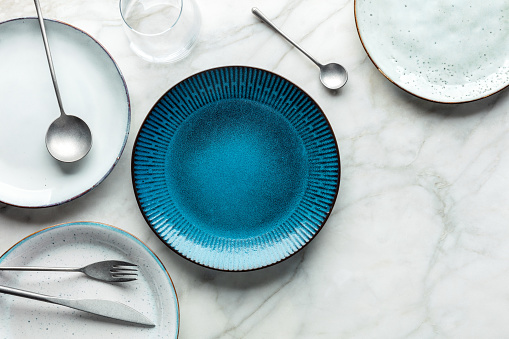 Modern tableware set with cutlery, a glass, and a vibrant blue plate, overhead flat lay shot with copy space. Trendy dinnerware on a marble background