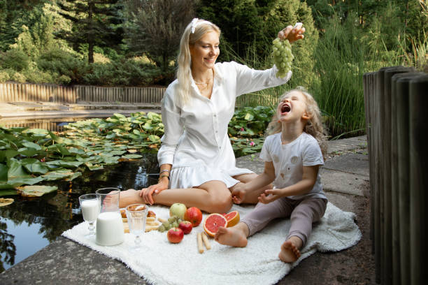 Funny family sitting on plaid with fruits, jug of milk on edge of pond. Young woman holding grapes over little girl. Portrait of funny family sitting on white plaid with fruits, jug of milk on edge of pond with green water lilies near fence in summer. Young woman holding grapes over little girl daughter. Picnic. feeding grapes stock pictures, royalty-free photos & images