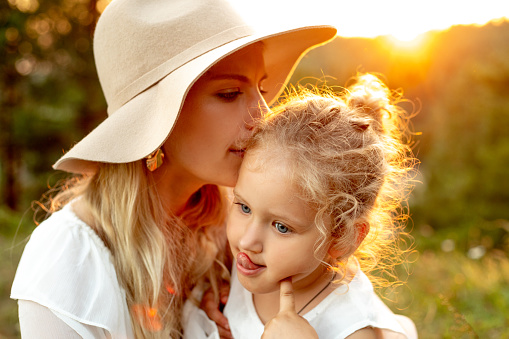 Portrait of loving family sitting in park forest illumined by sunset in summer. Young woman mother wearing beige hat, kissing temple of little girl daughter showing tongue, grimacing. Relationship.