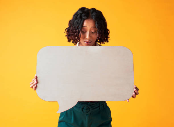 istock Woman holding an empty speech bubble board with copy space for a marketing poster in a studio. Girl with a blank advertising sign for an announcement. Lady with a small billboard design for branding. 1421009703