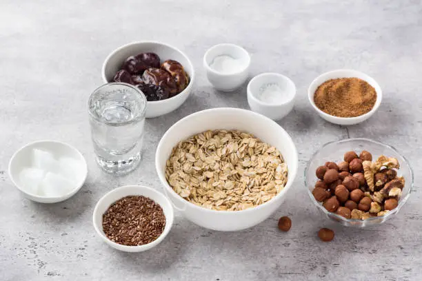 Ingredients for cooking healthy oatmeal cookies: oatmeal, flaxseed, nuts (hazelnuts and walnuts), coconut oil, water, dates, coconut sugar, salt and baking powder on gray textured background, top view