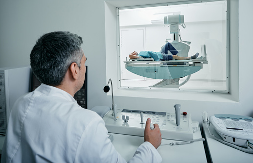 Radiologist controls X-ray process sitting behind protective window in his radiologists office while patient lying on bed of X-ray machine in radiographic imaging room