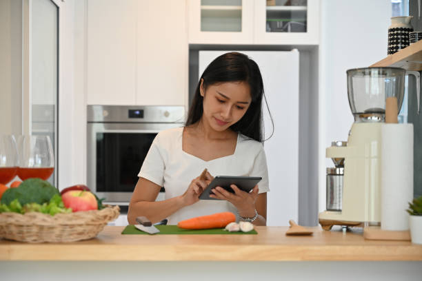 istock Young vegetarian woman 20s using digital tablet for looking  for recipe or learning cooking online with vegetables in kitchen at home, Domestic life and Healthy lifestyle concept. 1421015837
