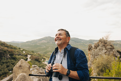 Happy senior man looking at the view while standing on a hilltop with binoculars. Cheerful elderly man enjoying a leisurely hike outdoors. Mature man enjoying recreational activities after retirement.