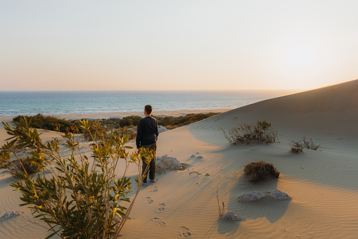Male explorer walking at the scenic sand dunes with a view of the Mediterranean sea during bright sunset in Patara, Antalya province
