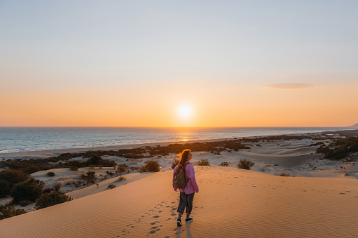 Young female backpacker in a pink sweatshirt walking at the scenic sand dunes with a view of the Mediterranean sea during bright sunset in Patara, Antalya province