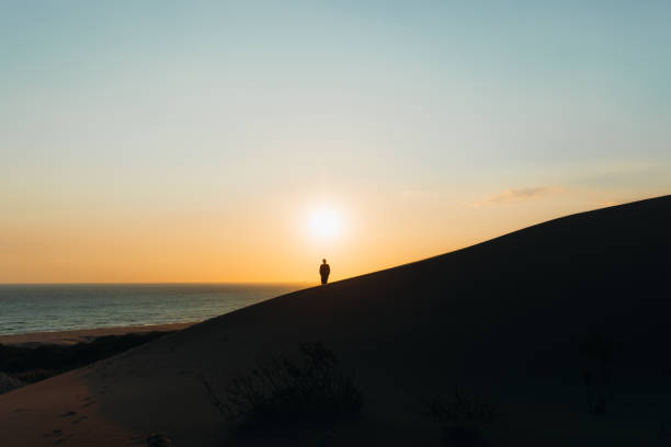 siluetta di un viaggiatore uomo che contempla il tramonto panoramico sopra le dune di sabbia in riva al mare in turchia - majestic landscape arid climate beach foto e immagini stock