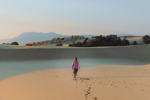 Young female backpacker in a pink sweatshirt walking at the scenic sand dunes with a view of the Mediterranean sea during bright sunset in Patara, Antalya province