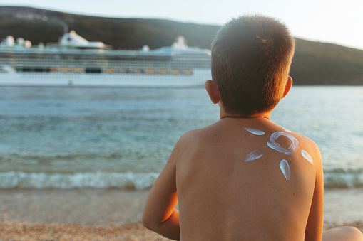 Shot of a beautiful little boy with a sun drawn on his shoulder with sunscreen at the beach, looking at the giant vessel. Protected against the sun.