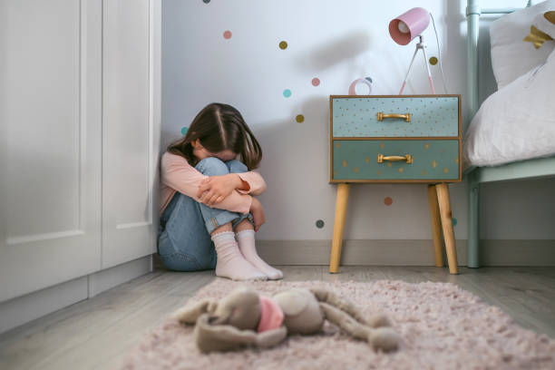 Sad little girl sitting on the floor of her bedroom with stuffed toy lying stock photo