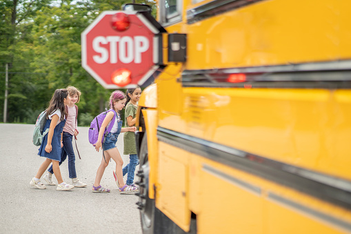A yellow school bus is parked with its flashing Stop sign folded out as a small group of students cross in front.  The students are dressed casually and have backpacks on as they make their way to board the bus.